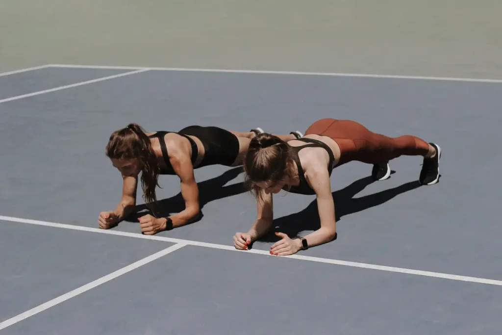 Two women in sportswear holding a plank position on an outdoor court, focusing on core strength and endurance.