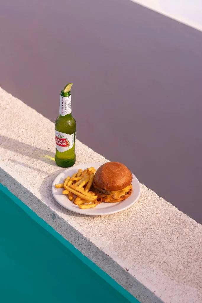 Plate with cheeseburger and fries next to a beer bottle by the poolside.