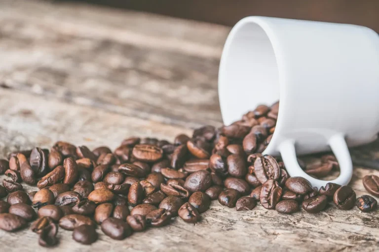 Roasted coffee beans spilling out of a white cup onto a rustic wooden surface.