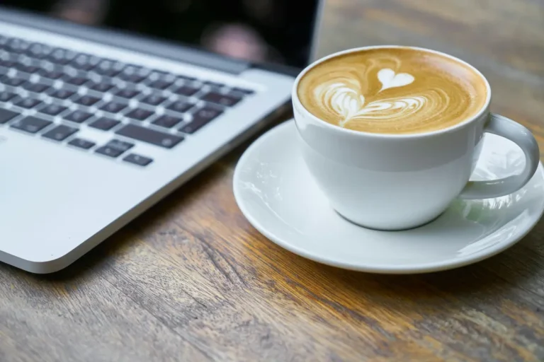 A cup of latte with heart-shaped foam art beside a laptop on a wooden table, symbolizing focus and productivity.