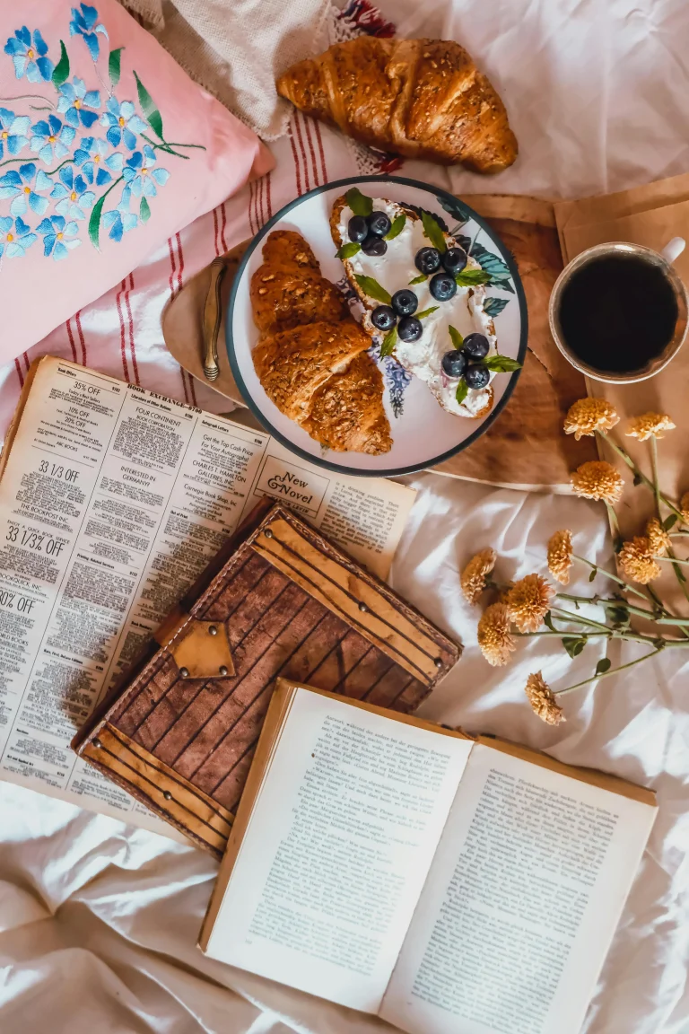 Flatlay of a cozy morning scene with croissants, coffee, books, and flowers on a bed