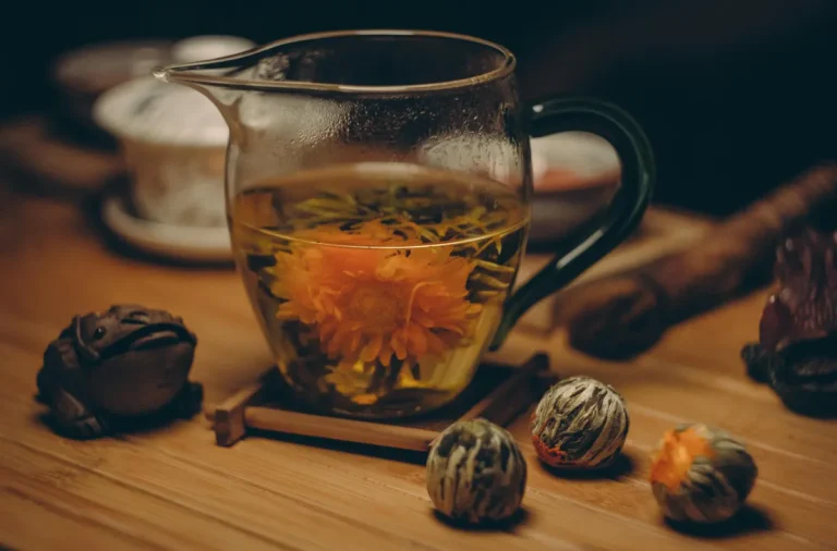 Glass teapot with blooming flower tea on a wooden surface, surrounded by dried tea buds and teaware.