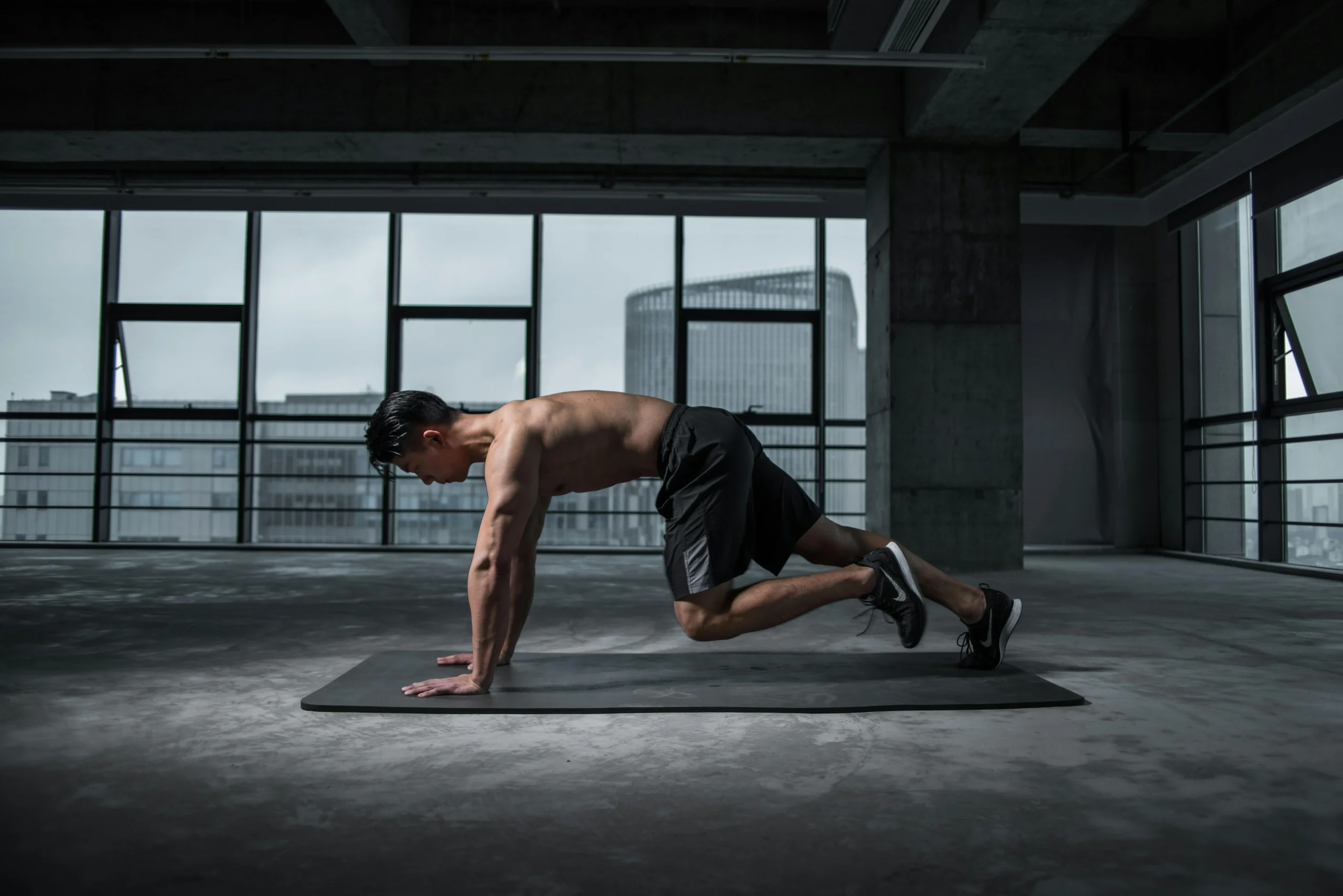 Shirtless man performing mountain climbers on a yoga mat in a gym with large windows