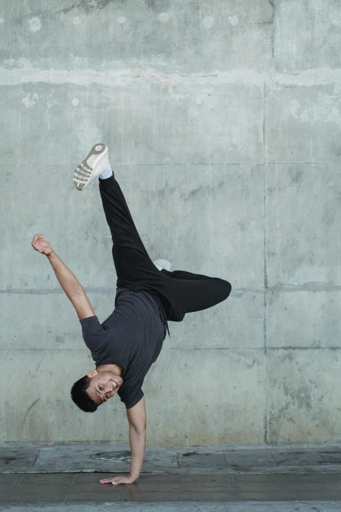 A man doing a one-handed handstand in casual athletic wear against a concrete wall, showcasing strength and balance