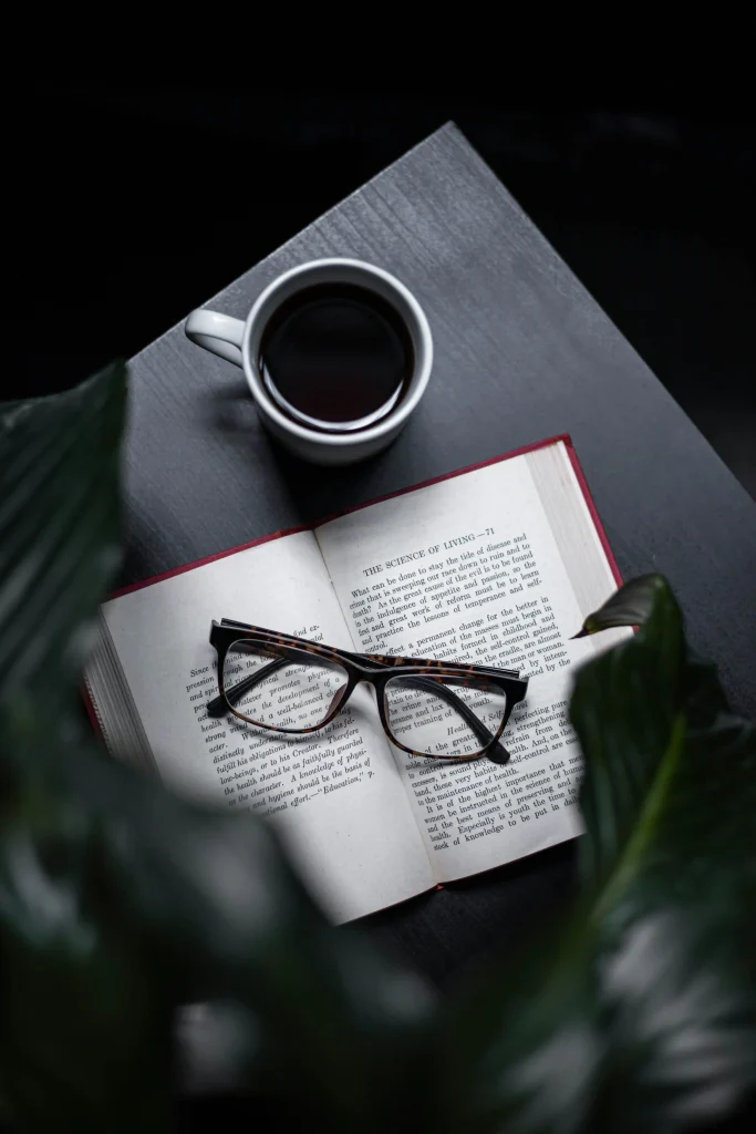 An open book with glasses resting on its pages next to a cup of black coffee on a dark table, surrounded by greenery.