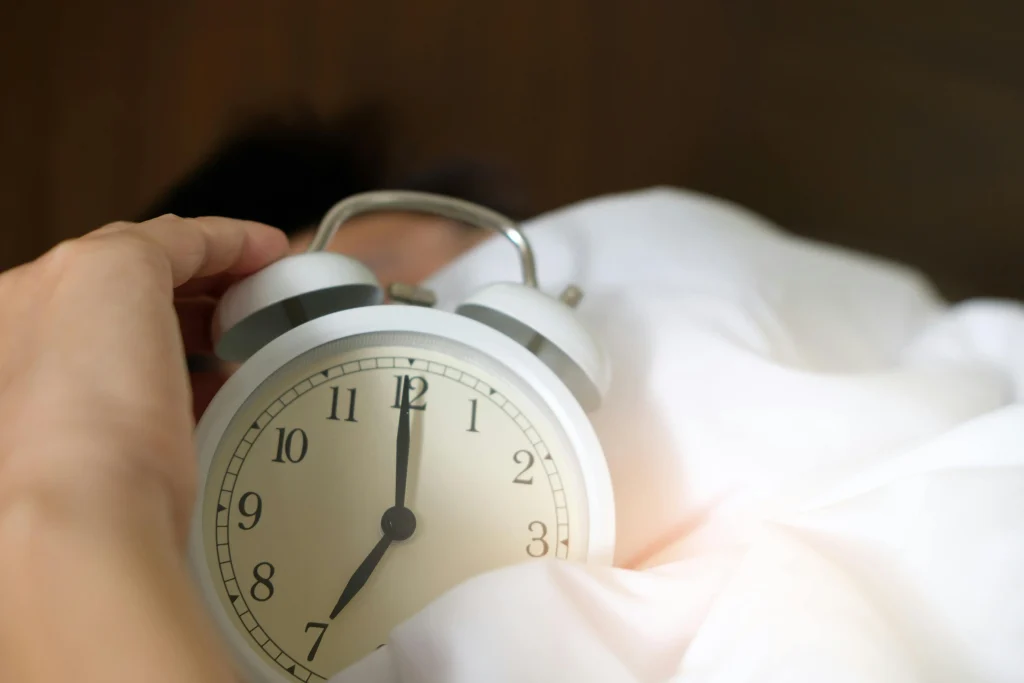 Person holding a white alarm clock in bed while waking up in the morning