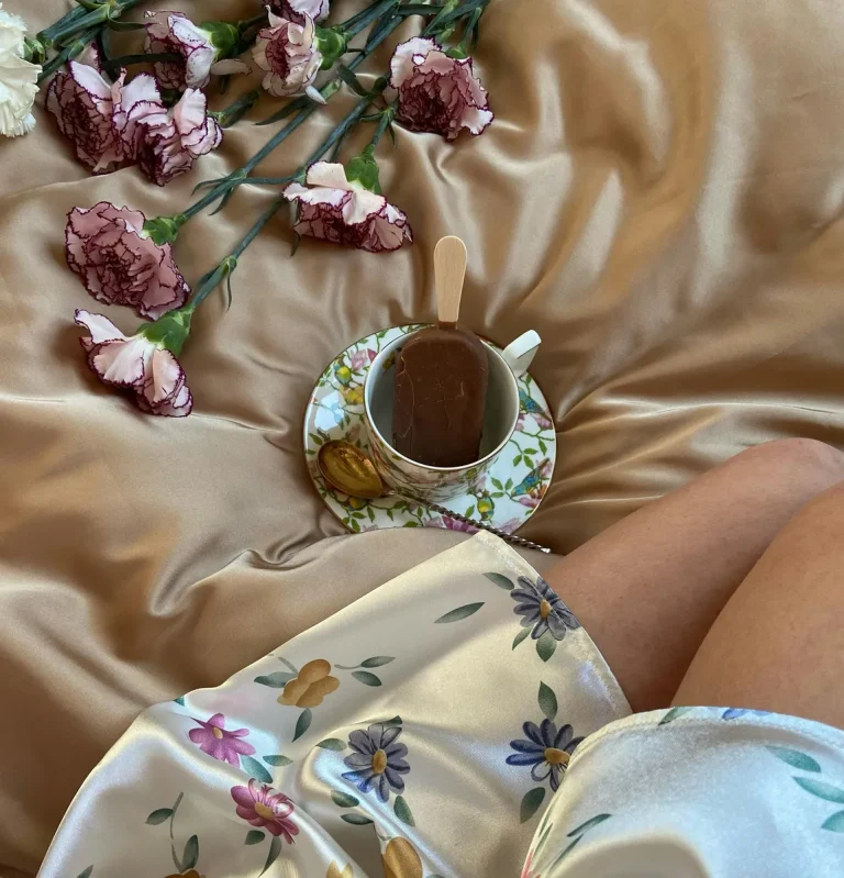 Cup of coffee with chocolate on top placed on a floral saucer with golden spoon, surrounded by pink and white flowers on silk bedding.