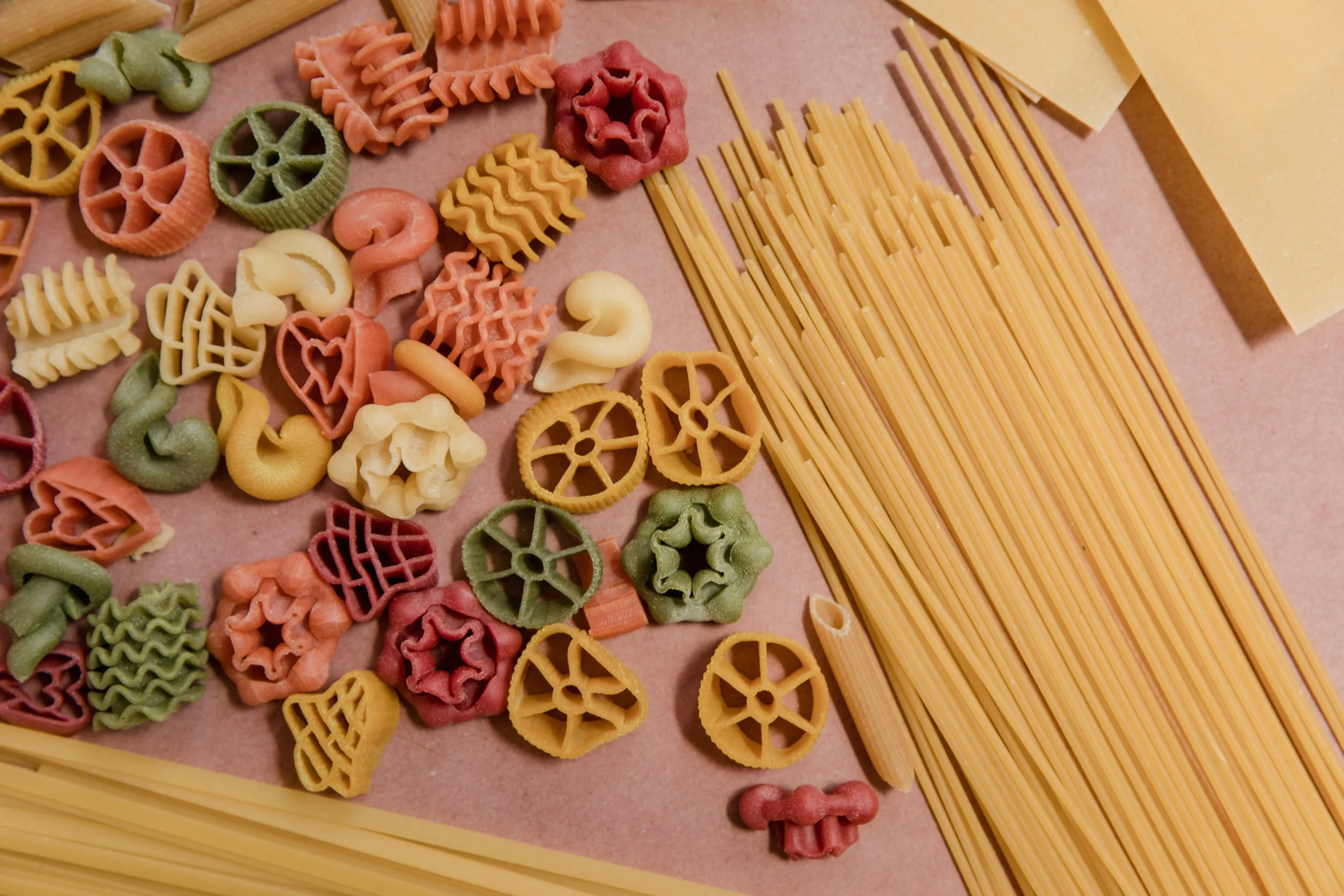 Colorful uncooked pasta including spirals, wheels, shells, spaghetti, and lasagna sheets arranged on a flat surface