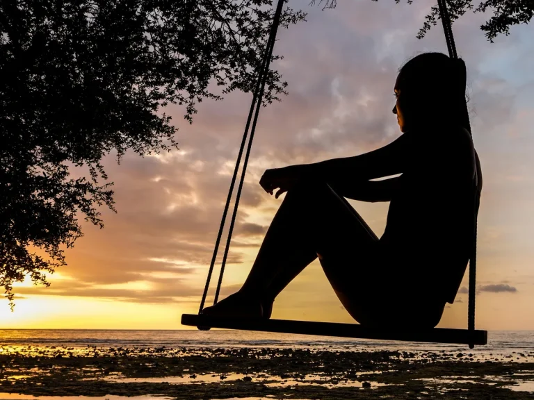 Silhouette of a woman sitting on a swing by the sea, watching the sunset with calm reflection and mindfulness.