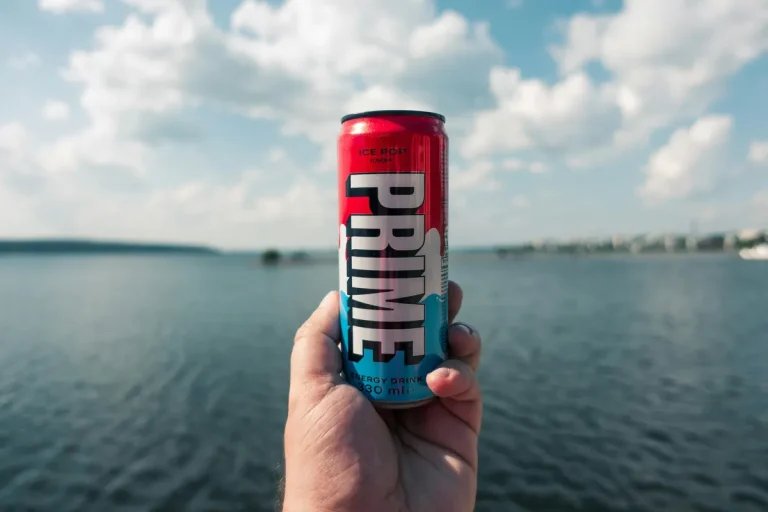 Hand holding a Prime energy drink can with a lake and cloudy sky in the background.