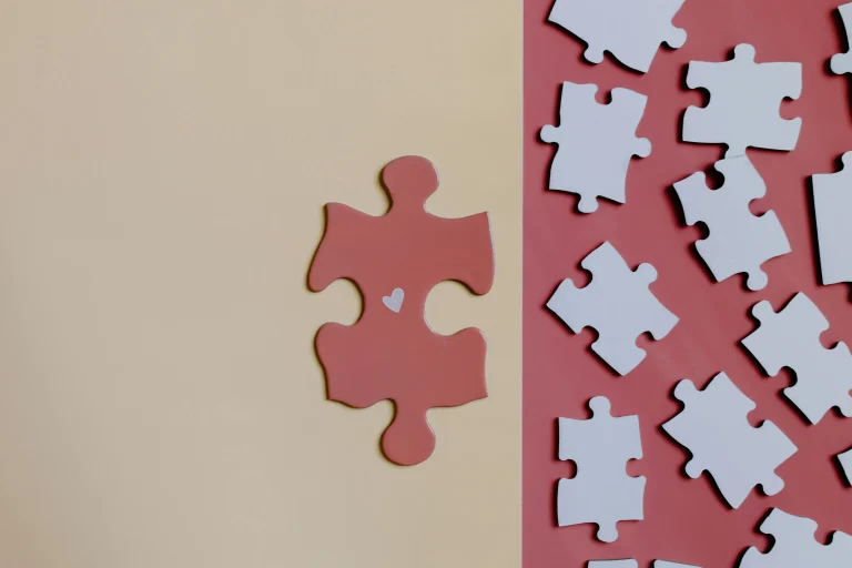 A pink puzzle piece with a small heart in the center placed beside white puzzle pieces on a pastel background