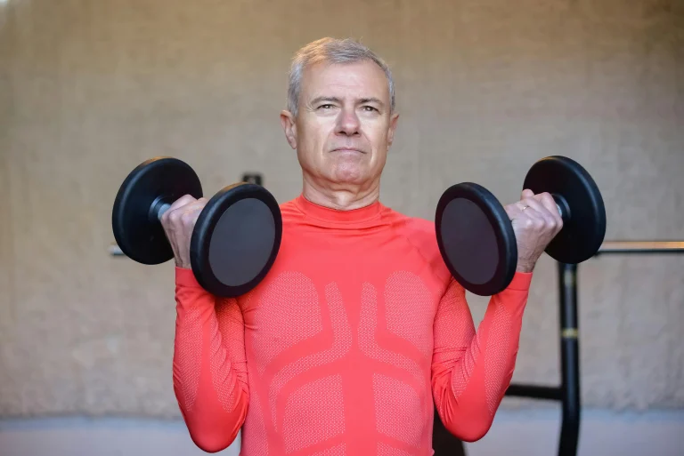 Older man in a red sports shirt lifting dumbbells indoors during a workout