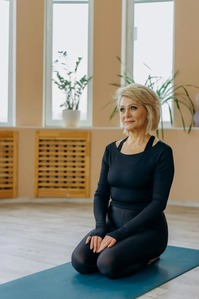 Older woman in black activewear sitting on a yoga mat in a calm studio practicing mindfulness and yoga