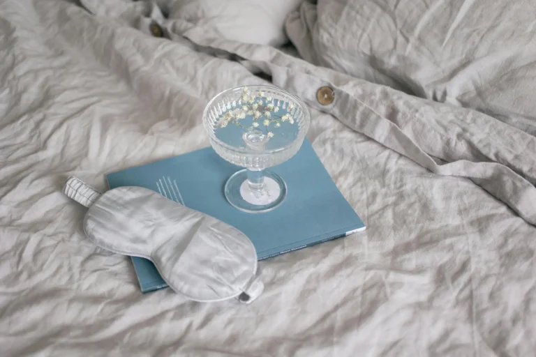 White sleep mask, blue book, and glass on a bed with neutral bedding.