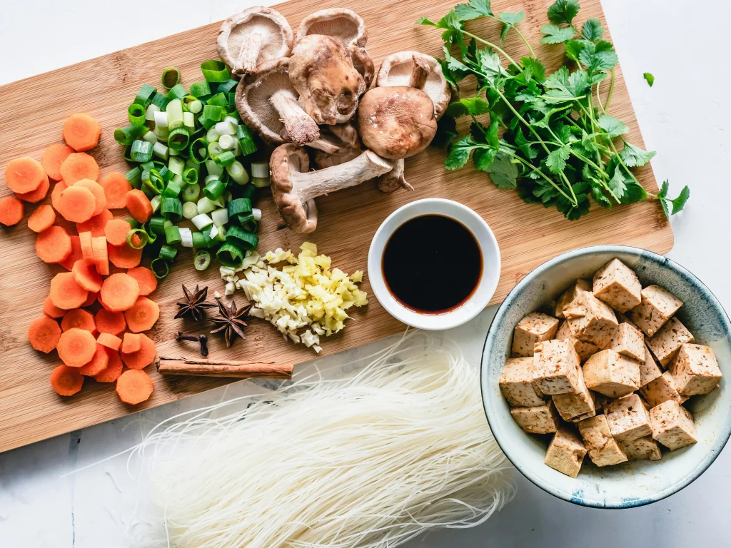 Fresh tofu cubes, carrots, green onions, mushrooms, herbs, and noodles arranged on a wooden board for a healthy meal