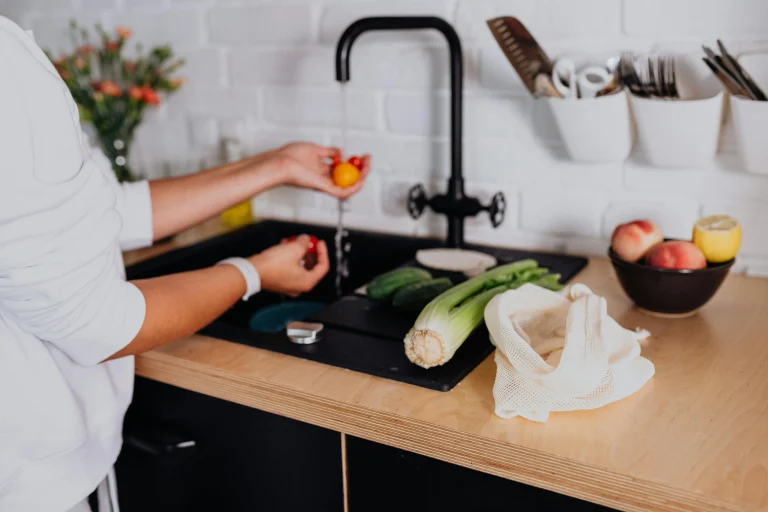 Person washing vegetables under running water in a kitchen sink with celery, cucumbers, and fruits on the counter