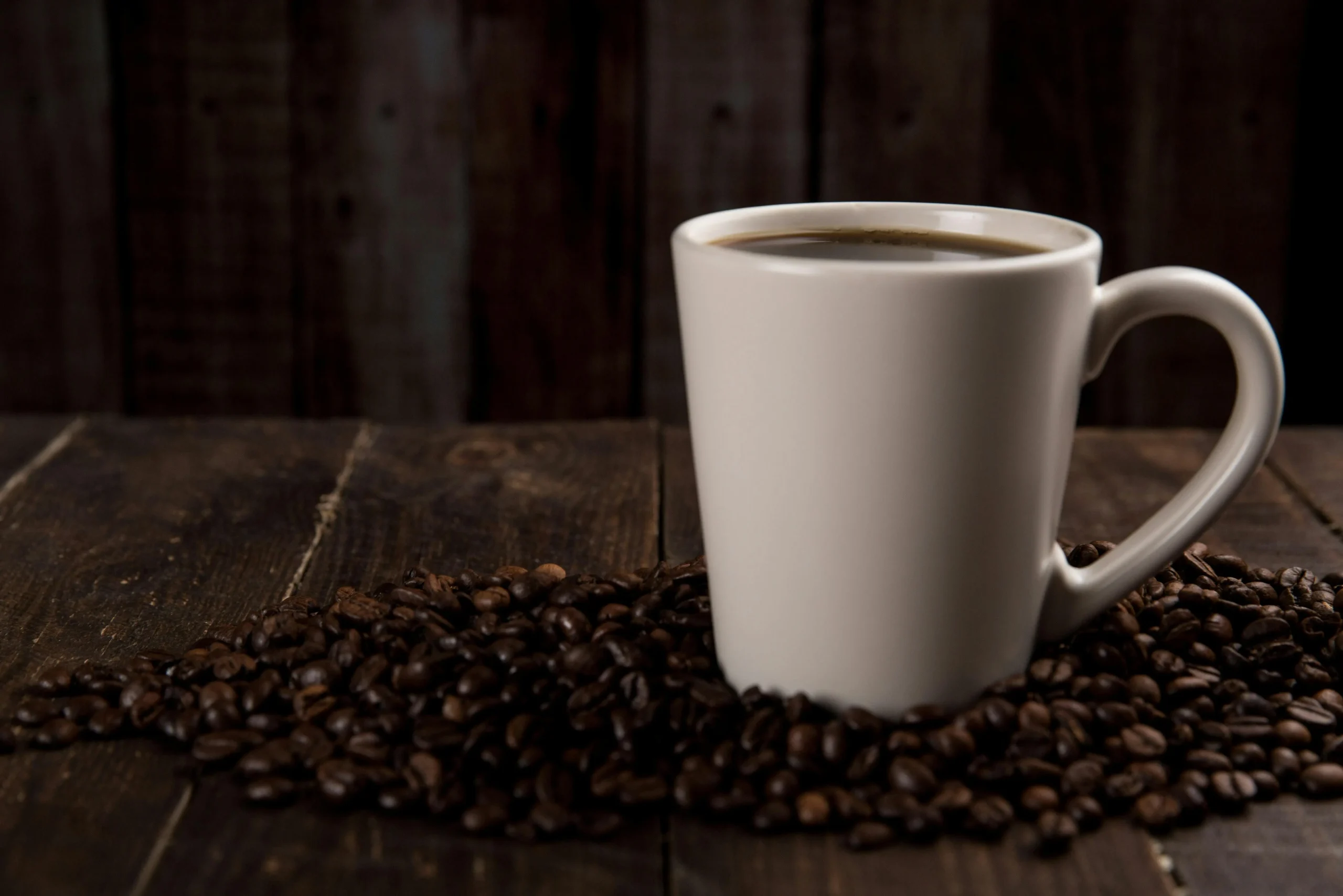 White ceramic coffee cup filled with hot coffee surrounded by roasted coffee beans on a wooden table