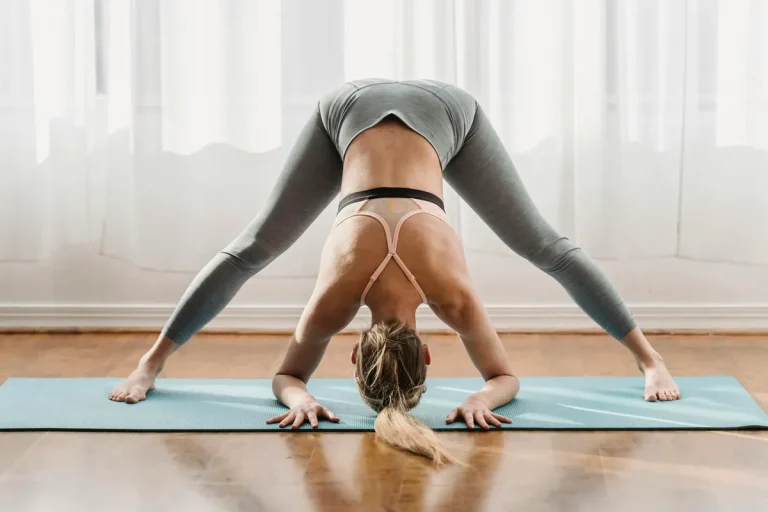 Woman practicing wide-legged forward bend pose on a yoga mat indoors, with head lowered and hands on the floor