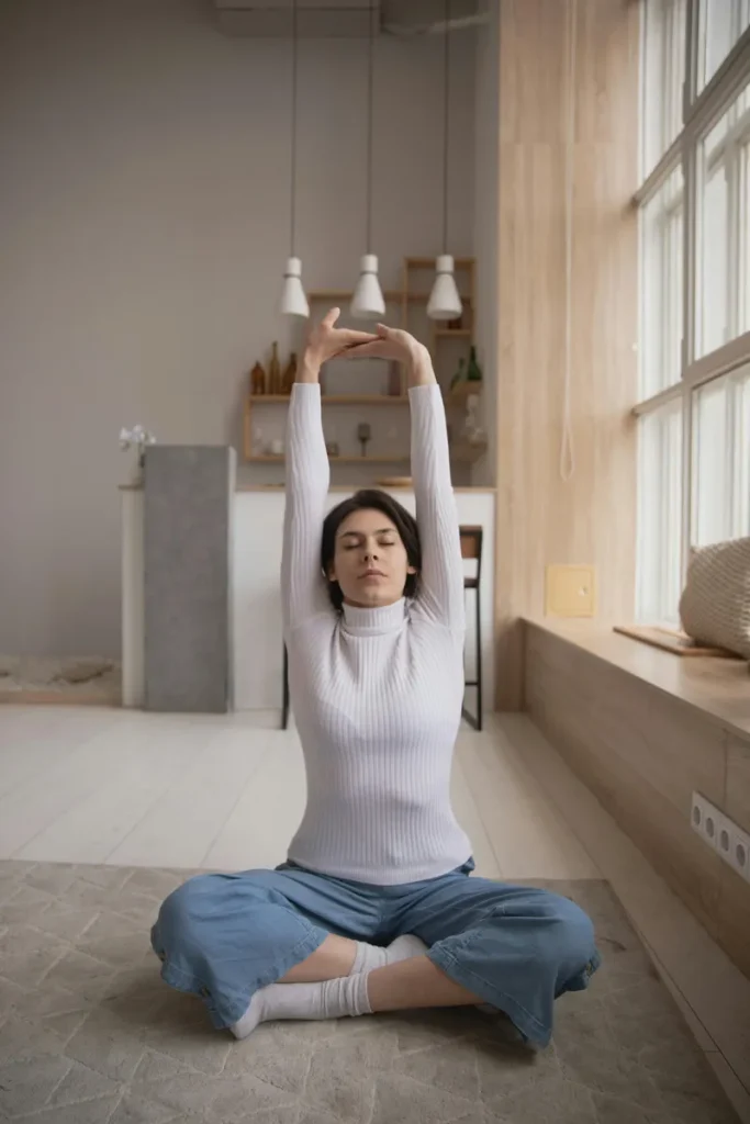 Young woman sitting cross-legged on the floor with eyes closed, stretching arms overhead in a calm home setting