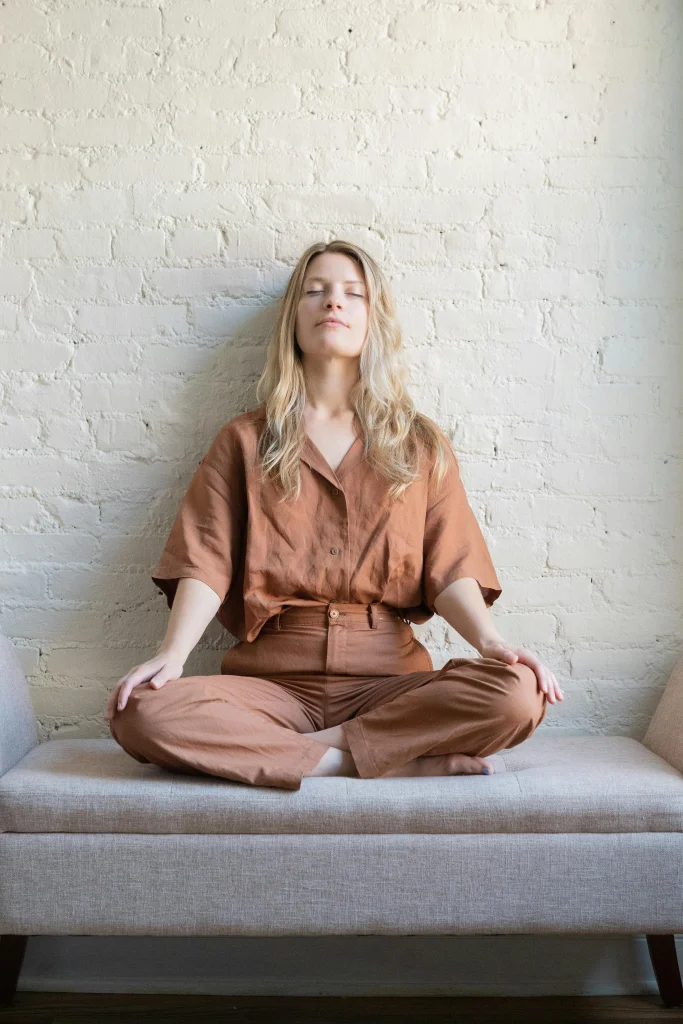 Young woman sitting cross-legged on a sofa with eyes closed, meditating in front of a white brick wall