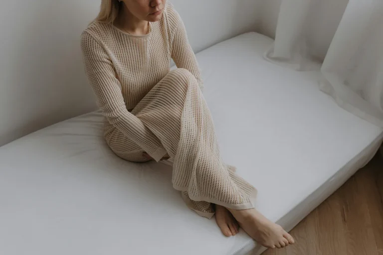 Woman in a beige textured outfit sitting thoughtfully on a simple white bed in a minimalist room with natural light.