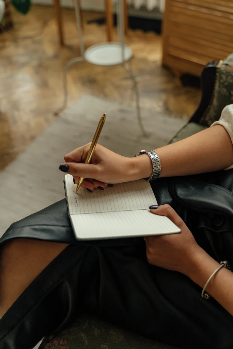 Close-up of a woman’s hands writing in a lined notebook with a golden pen while sitting indoors