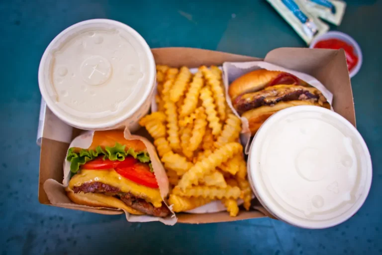 Takeaway box containing two cheeseburgers with lettuce and tomato, crinkle-cut fries, and two covered drinks.