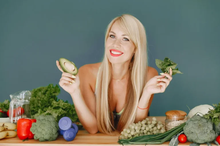 Smiling woman holding an avocado and leafy greens surrounded by fresh fruits, vegetables, and dumbbells on a table.