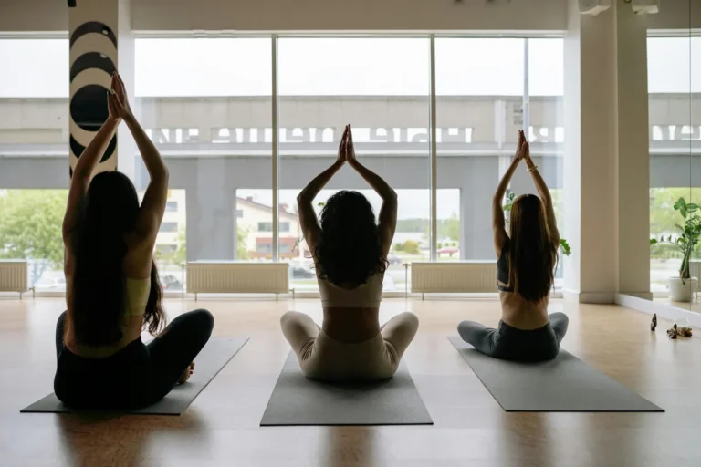 Three people seated in cross-legged prayer pose on yoga mats in studio with large windows and urban backdrop
