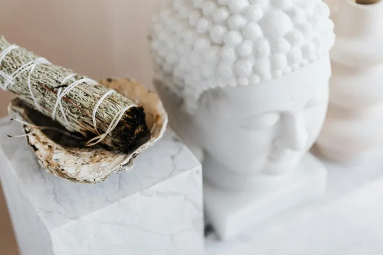A white Buddha head statue beside a burning sage smudge stick in a shell on a marble surface.